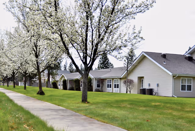 Single-story senior living buildings with blossoming trees lining a sidewalk and a well-kept green lawn.