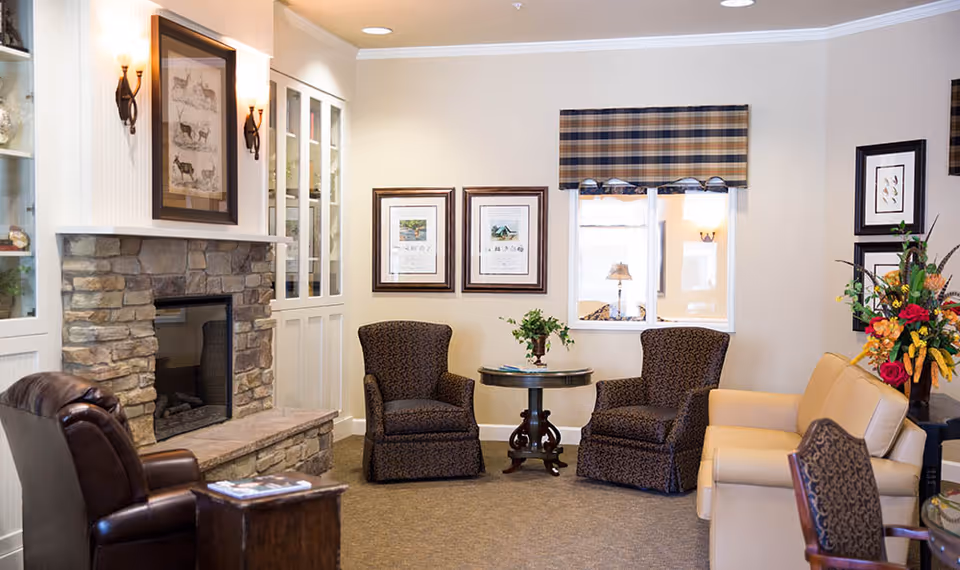 A cozy living room area with a stone fireplace on the left, two patterned armchairs around a small round wooden table, a beige sofa, and a leather chair. The walls are decorated with framed pictures and a window with a plaid valance. There is a vase with colorful flowers on a side table to the right.