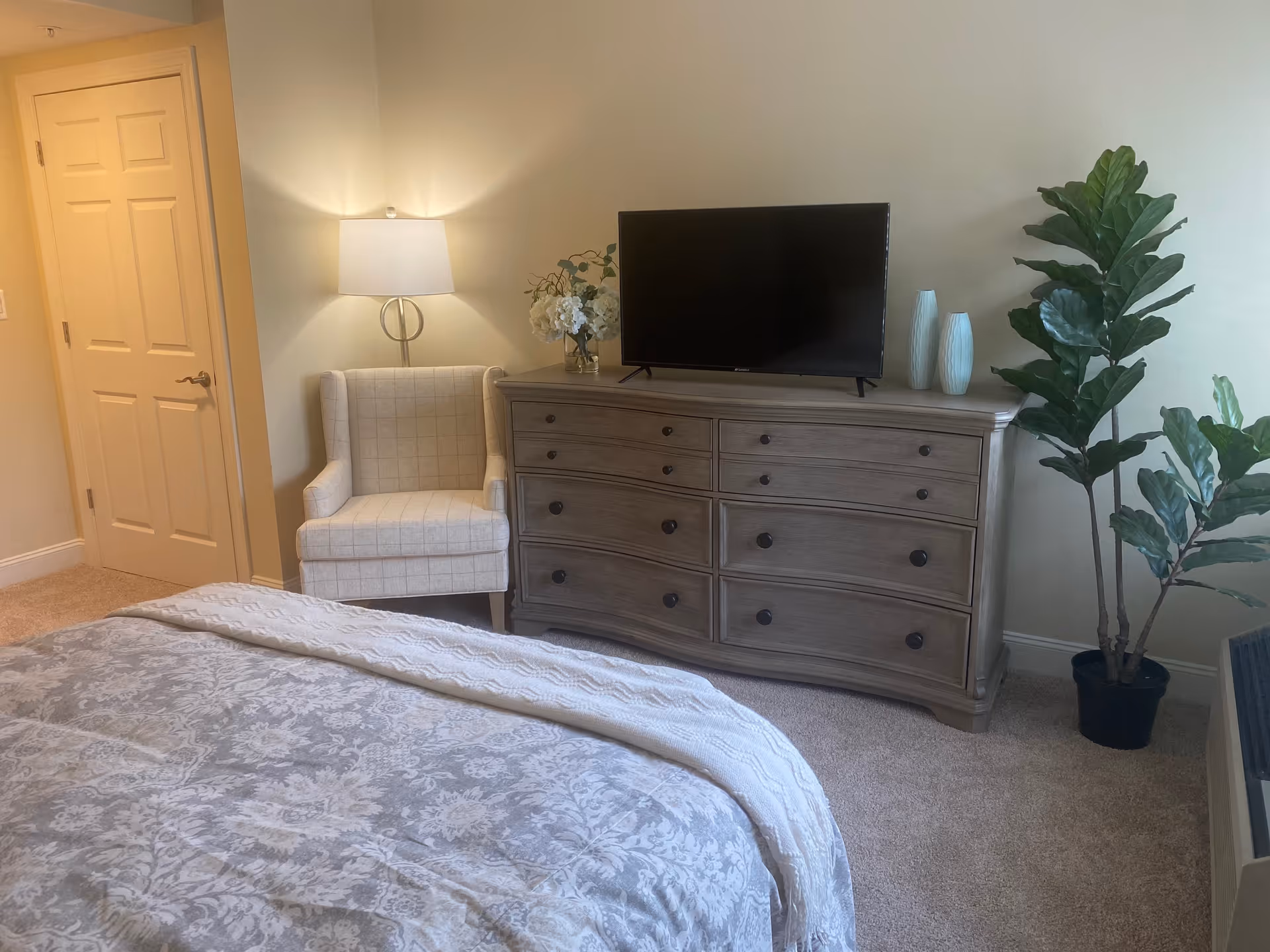 A bedroom with a patterned bedspread in the foreground, a beige armchair with a floor lamp behind it, a wooden dresser with a flat-screen TV on top, decorative vases, a flower arrangement, and a tall potted plant next to the dresser. A closed door is visible on the left side of the room.