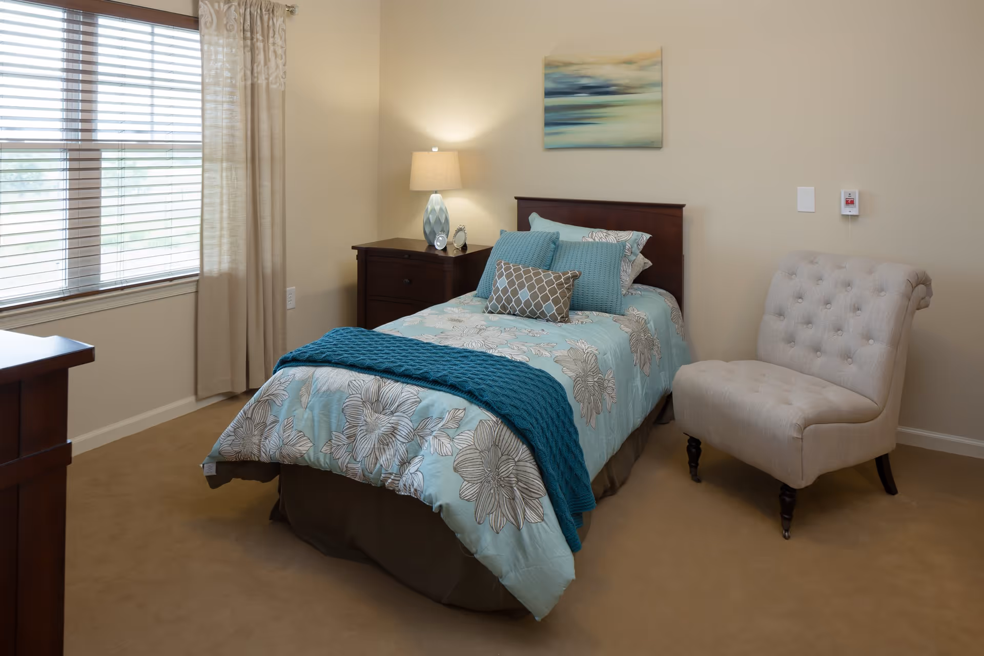 A cozy bedroom with a single bed covered in a floral patterned blue comforter and multiple pillows. Next to the bed is a dark wooden nightstand with a decorative lamp and small clock. A beige upholstered chair is placed near the bed. The room has beige walls, a window with blinds and light curtains, and a piece of abstract art hanging above the bed.