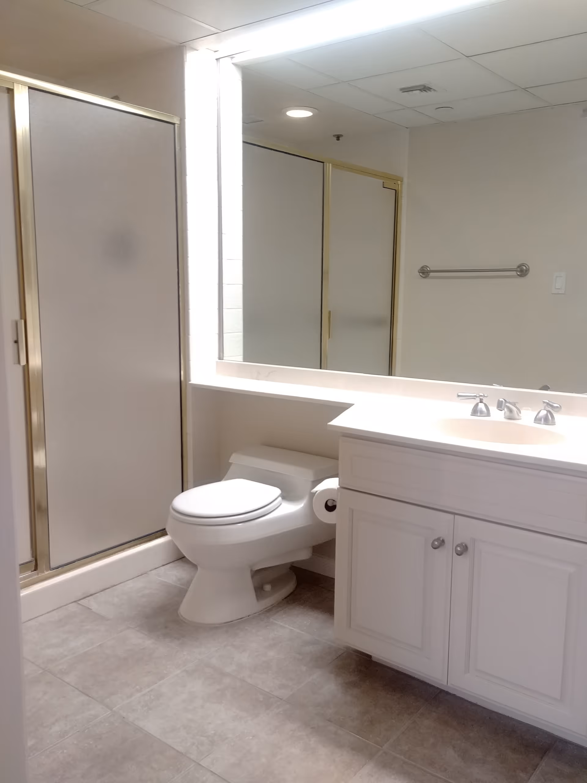 A clean bathroom featuring a white toilet, a white vanity with a sink and silver faucet, a large mirror above the vanity, and a shower with frosted glass doors and gold trim. The floor is tiled in a light gray color.