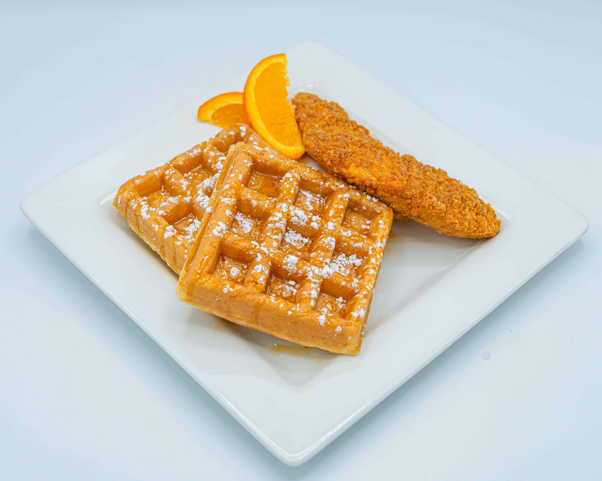 Two powdered waffles and a breaded chicken strip with orange slices on a white square plate.