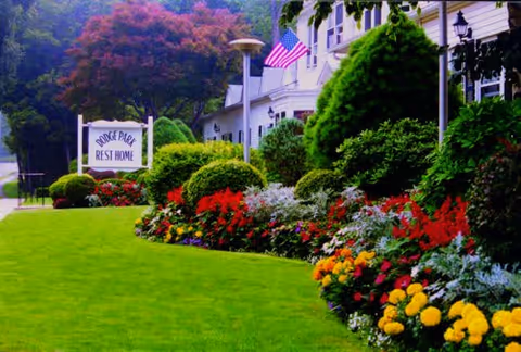 Well-maintained garden with colorful flowers and neatly trimmed bushes in front of a white building with an American flag. A sign reads 'Dodge Park Rest Home'.