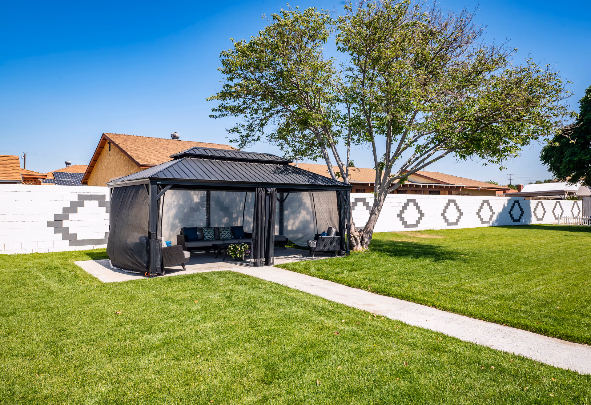 Outdoor area with a green lawn, a concrete pathway, a large tree, and a black gazebo with mesh sides containing outdoor seating furniture including chairs and a sofa. A white brick wall with geometric patterns is visible in the background under a clear blue sky.