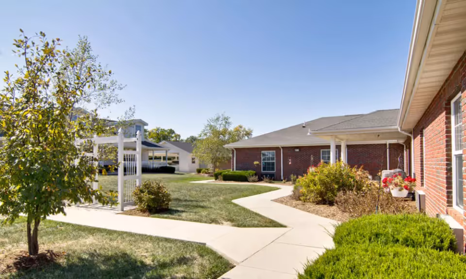 Sunny courtyard with paved walkways, lawn, shrubs, a white pergola, and single-story brick senior living buildings.