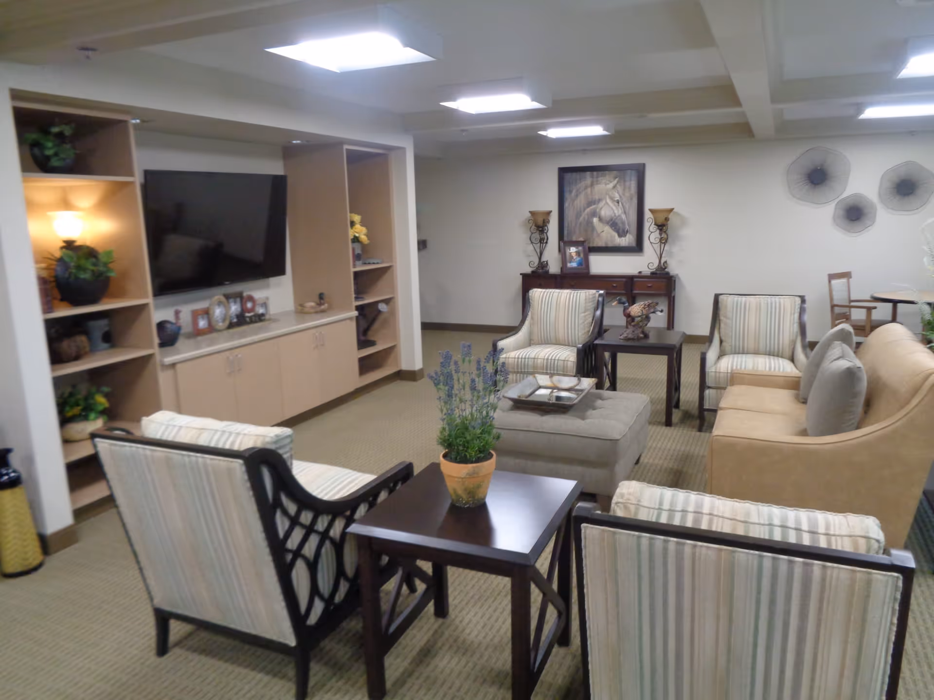 A cozy living room area in an assisted living center featuring a beige sofa, four striped armchairs, a central ottoman, and two dark wooden side tables with a potted plant on one. The room has built-in shelves with decorative items and a large flat-screen TV mounted on the wall. The walls are adorned with a framed horse picture and decorative wall art, with soft lighting from ceiling fixtures and table lamps.