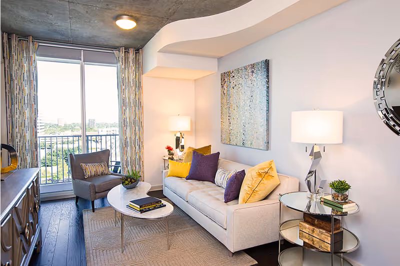 Bright modern living room with a sofa and colorful throw pillows, armchair, coffee table, lamps, and a balcony window.