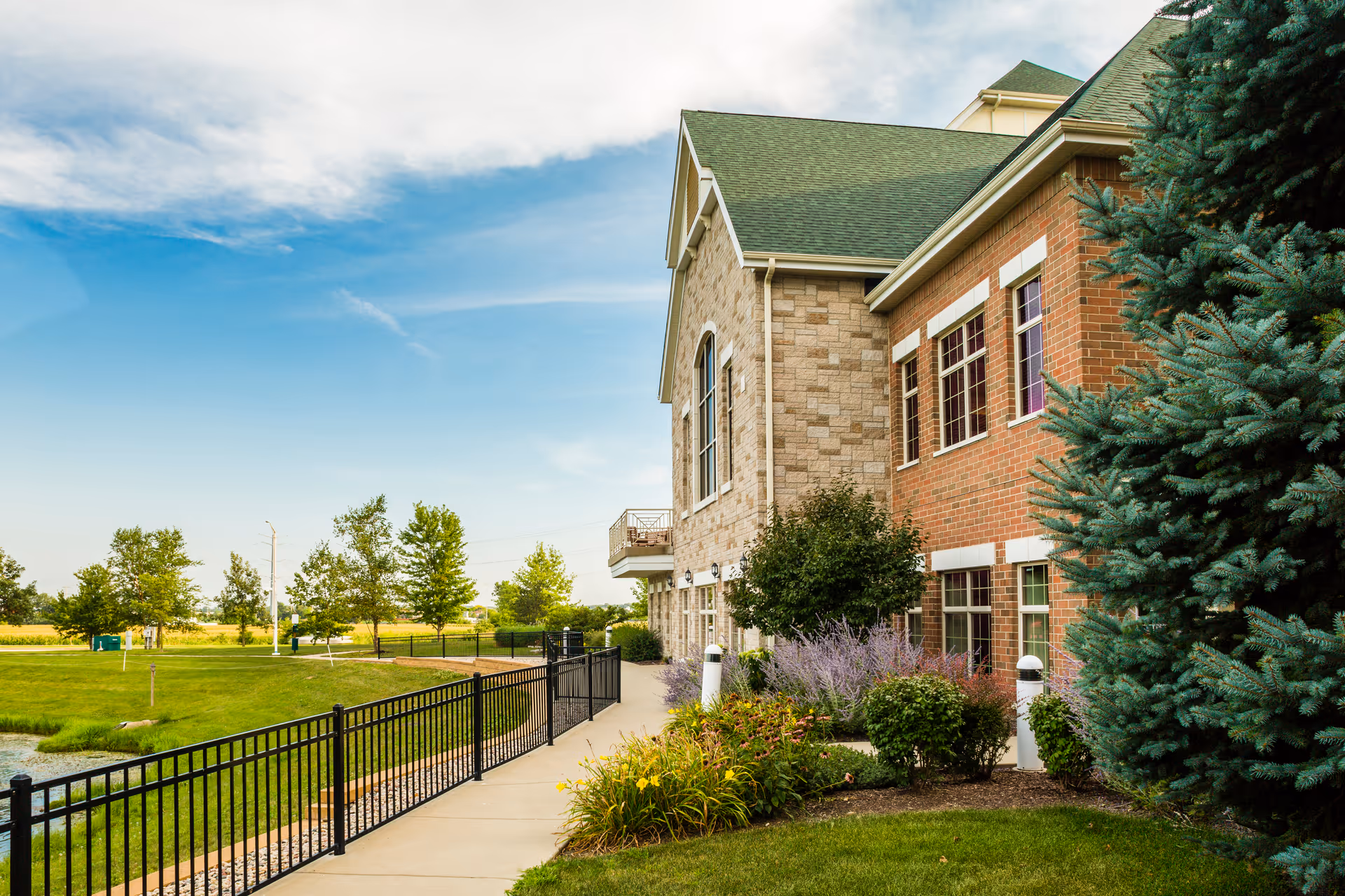 Exterior view of a senior living facility building with a mix of brick and stone facade, green roof, and multiple windows. There is a paved walkway with a black metal fence alongside landscaped greenery including bushes, flowers, and a large evergreen tree. The sky is partly cloudy with blue patches.
