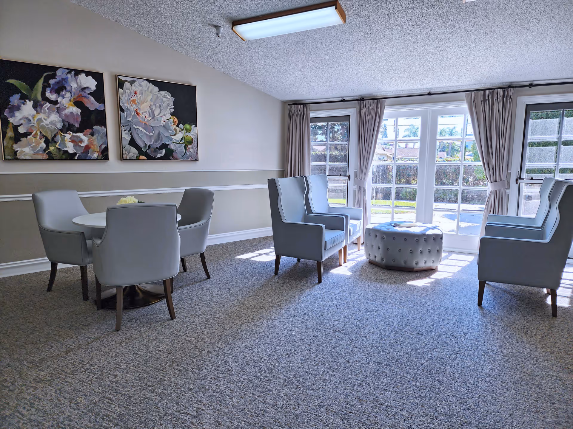A bright and airy sitting area with four light gray armchairs arranged around a round white table on the left and four matching armchairs arranged around a round tufted ottoman near large windows with beige curtains letting in natural light. Two floral paintings hang on the wall above the table.