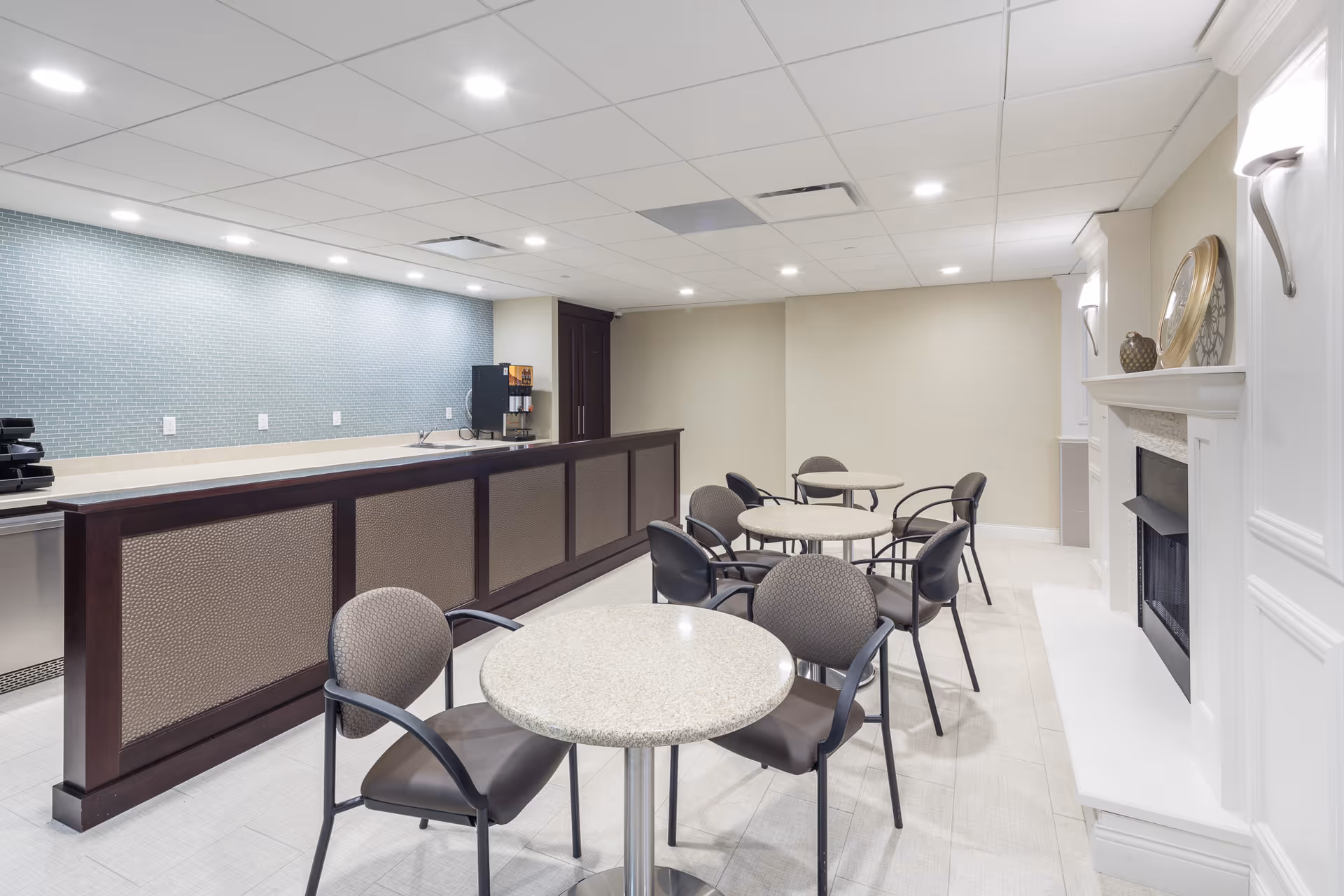 A clean and modern common area with round tables and chairs arranged for seating. There is a long counter with a coffee machine and a light blue tiled backsplash. The room features a white fireplace with decorative items on the mantel and soft lighting fixtures on the walls.