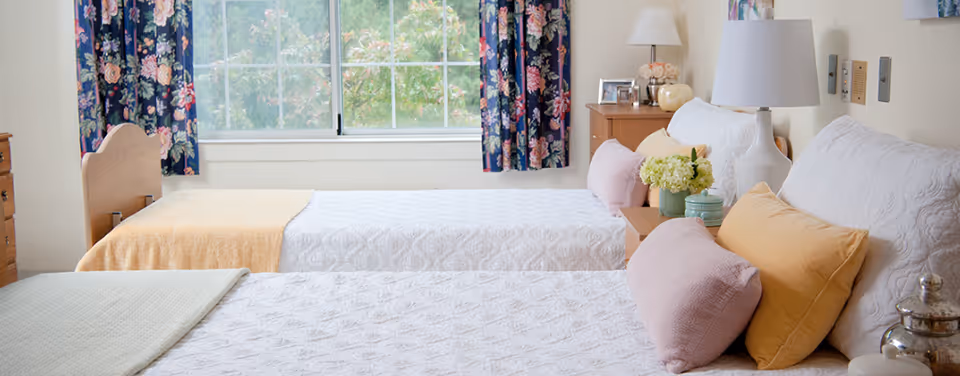 A bright bedroom with two twin beds covered in white quilts and pastel-colored pillows. A wooden nightstand between the beds holds a lamp, framed photo, and decorative items. Floral curtains frame a large window overlooking greenery outside.