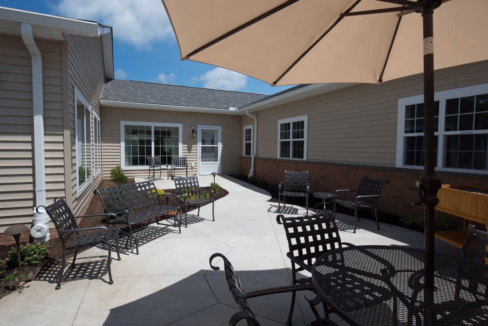 Outdoor patio area at Kemper House Highland Heights with metal chairs and tables, some shaded by a large umbrella, surrounded by beige siding buildings under a blue sky with some clouds.