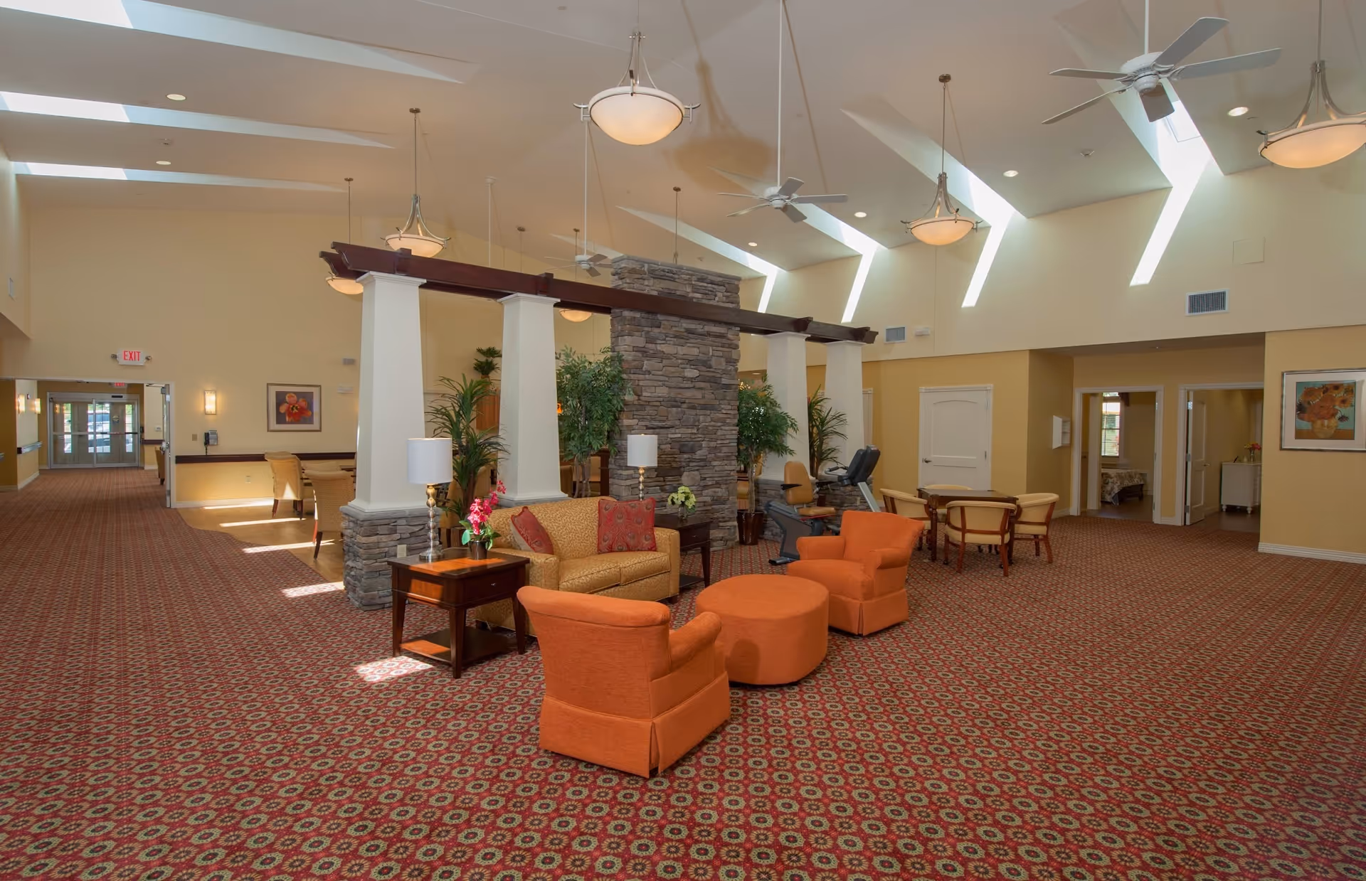 A spacious and well-lit common area with high ceilings and skylights. The room features a patterned carpet, orange armchairs, a matching ottoman, a beige sofa with red cushions, side tables with lamps, and several potted plants. There is a stone pillar in the center with wooden beams extending from it. In the background, there are additional seating areas and doorways leading to other rooms.