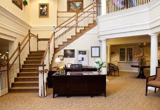 Interior view of a senior living facility lobby with a dark wooden reception desk in the center, a staircase with wooden handrails leading to an upper floor, framed artwork on the walls, and seating areas with chairs and tables. The space is well-lit and decorated with plants and lamps.