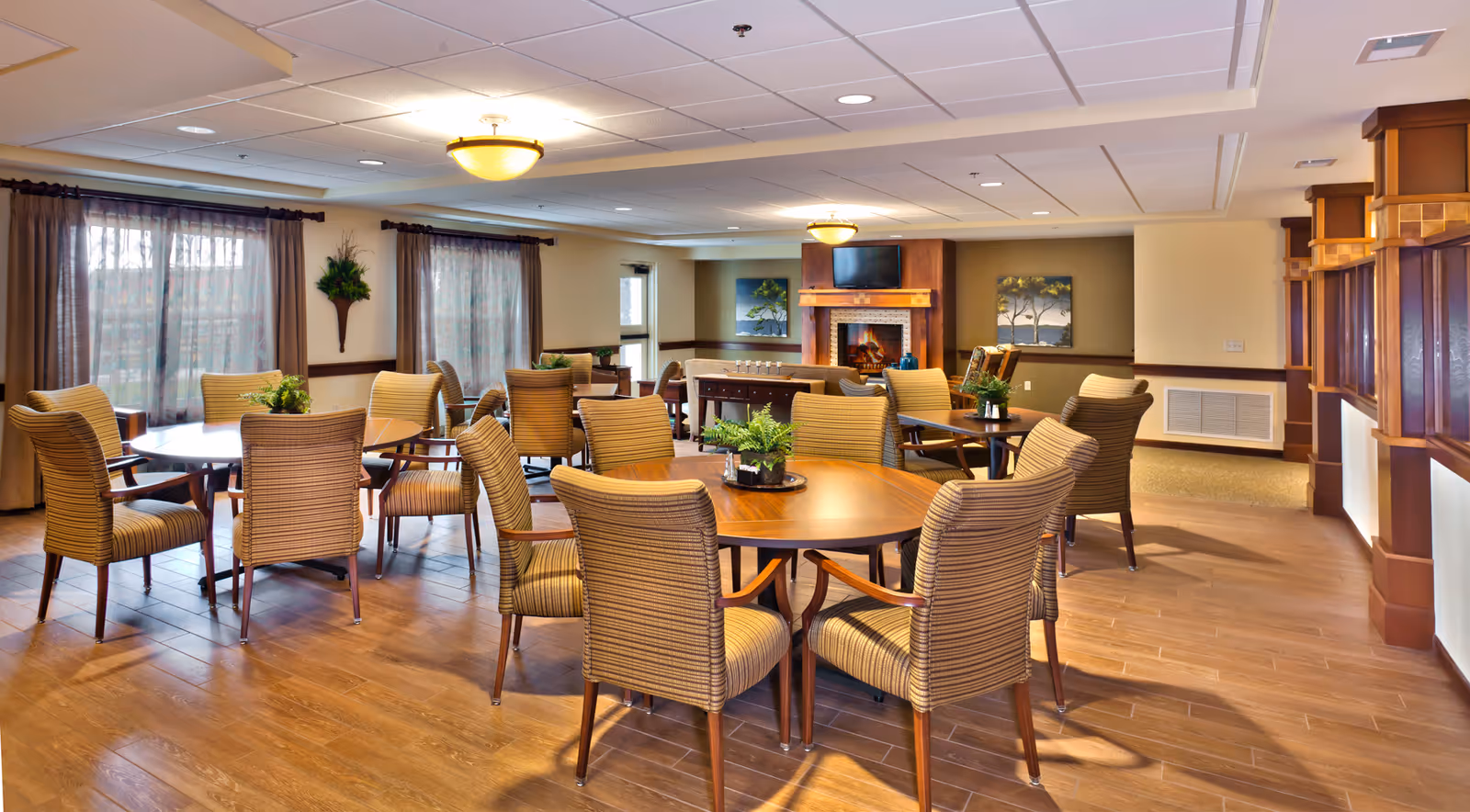 Bright communal dining/lounge area with round wooden tables, upholstered chairs, a fireplace and TV.