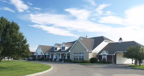 Exterior view of a single-story senior living facility building with a curved driveway, manicured lawn, and trees under a partly cloudy blue sky.
