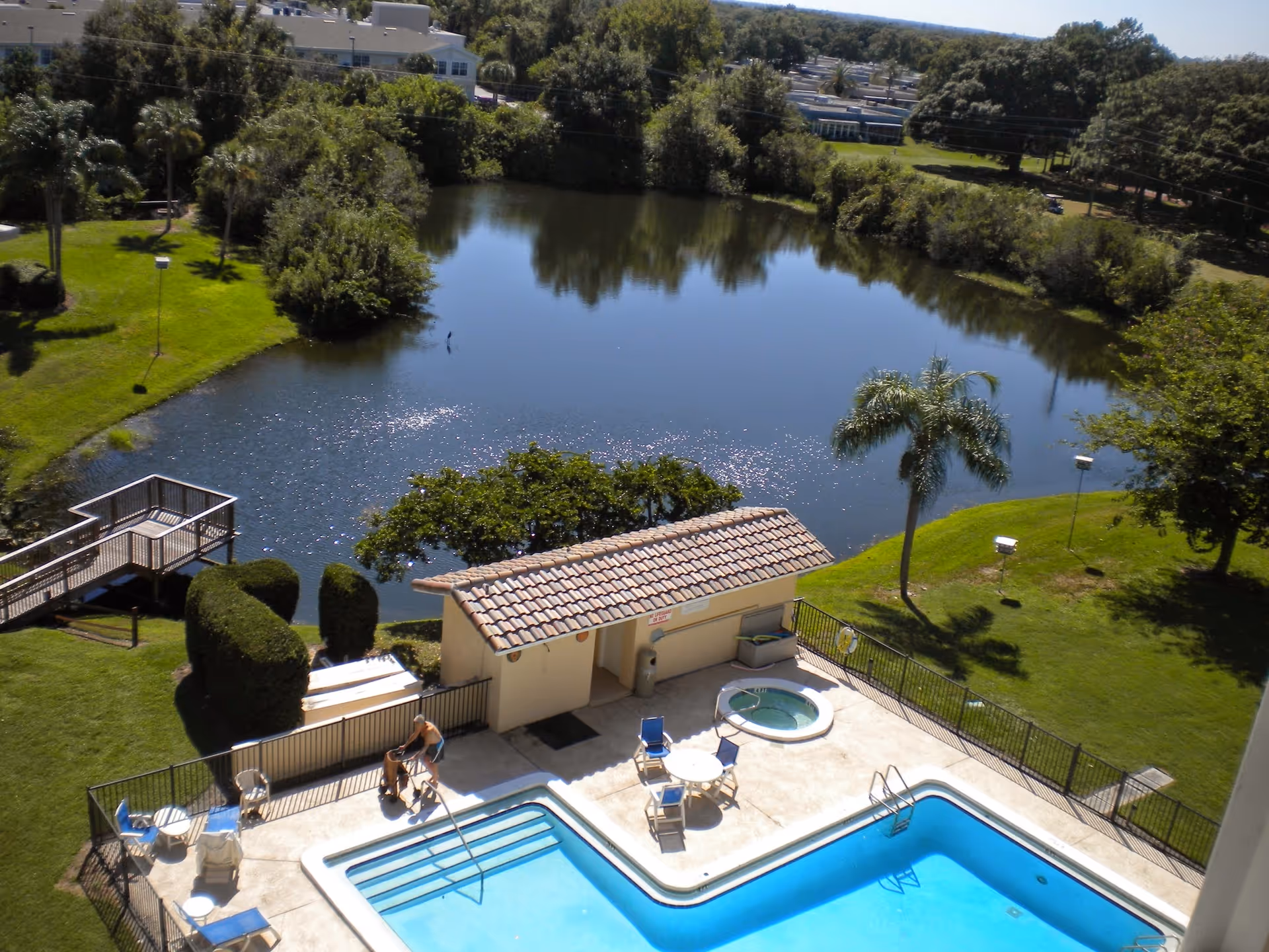 Aerial view of an outdoor pool area with a small hot tub, several chairs and tables, and a tiled roof structure next to a pond surrounded by green grass, trees, and bushes. A person is seen near the pool area, and there is a wooden dock extending into the pond.