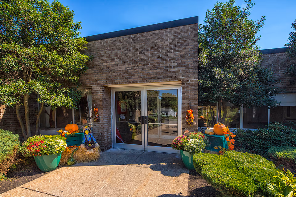 Brick building entrance with glass double doors flanked by pumpkins, fall planters, and shrubs.