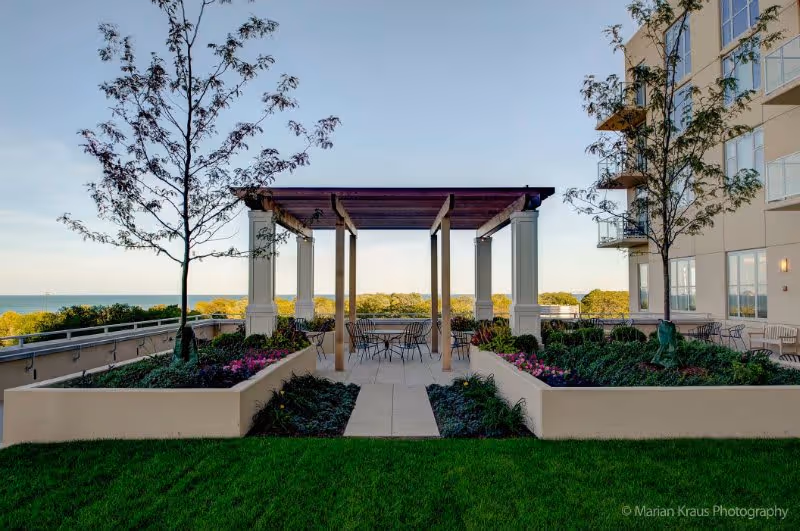 Outdoor patio area with a pergola supported by four columns, surrounded by raised garden beds with flowers and small trees. There are metal chairs and tables under the pergola and on the patio. A multi-story building is visible on the right side, and trees and a body of water can be seen in the background under a clear sky.