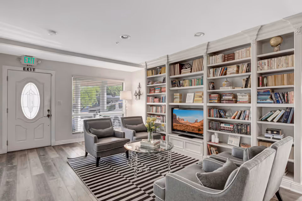 A cozy living room with a white front door featuring an oval glass window, a large window with blinds, two gray armchairs, a gray sofa, a glass coffee table on a black and white striped rug, and a built-in white bookshelf filled with books and a flat-screen TV displaying a desert landscape.