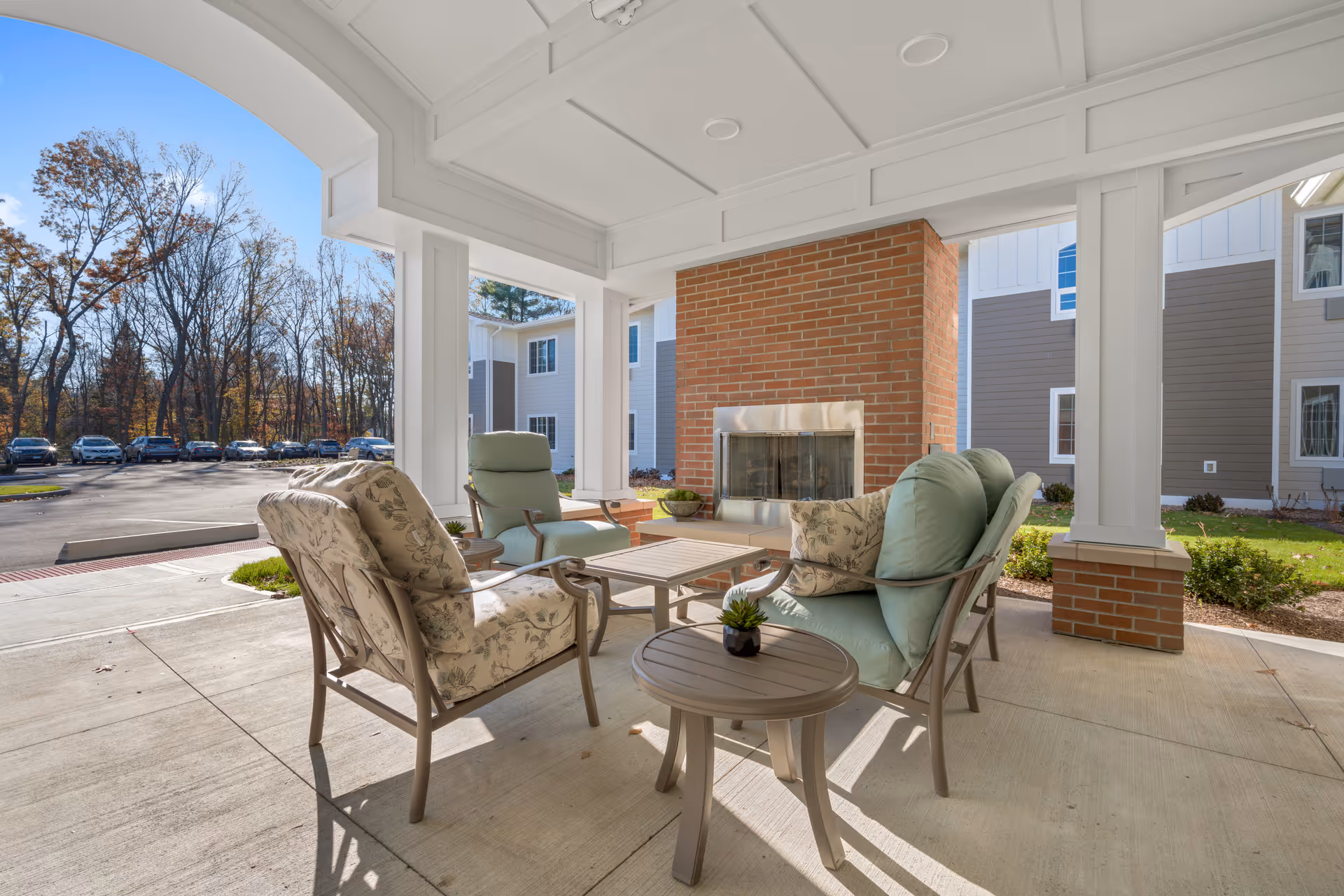 Outdoor covered seating area with cushioned chairs and a small table arranged around a brick fireplace, with a view of a parking lot and trees in the background at The Landing of North Haven.