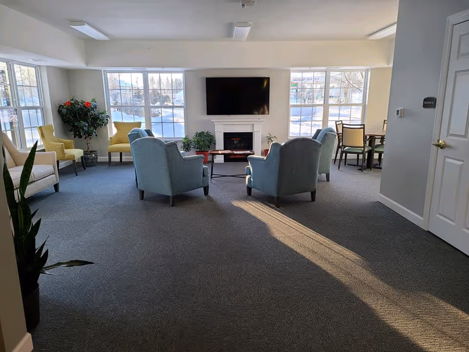 A bright and spacious living room area with several armchairs arranged around a coffee table in front of a wall-mounted flat screen TV and an electric fireplace. Large windows let in natural light and offer a view of a snowy outdoor scene. There are also some potted plants and a dining table with chairs in the background.