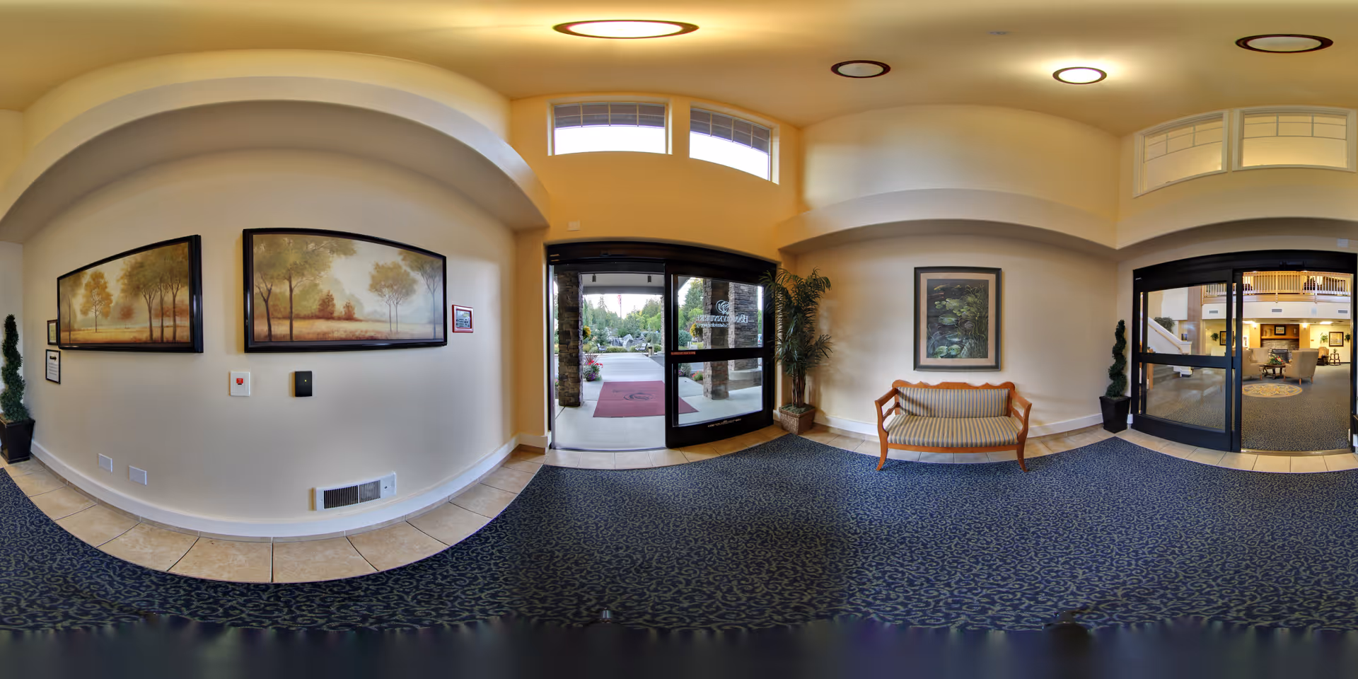 Interior view of the entrance lobby of Bonaventure of Lacey, featuring double glass doors leading outside, a cushioned wooden bench against the wall, framed artwork, potted plants, and a carpeted floor with a patterned design.