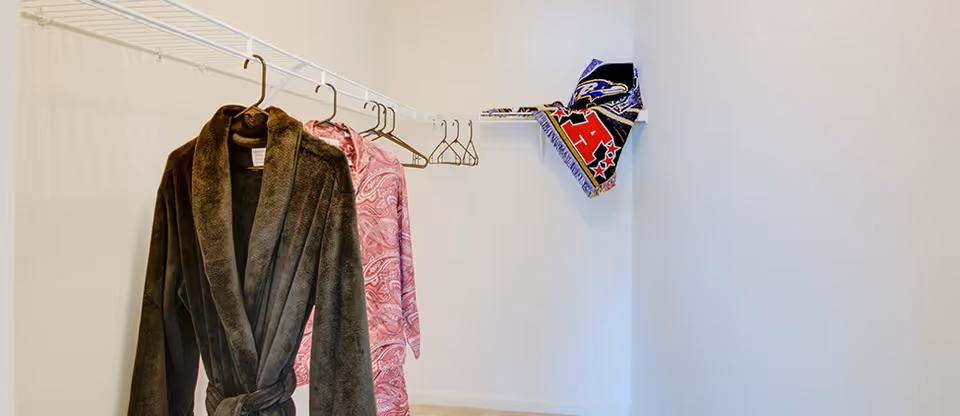 A walk-in closet with white walls and a beige carpet. There is a white wire shelf with several hangers, two of which hold a brown robe and a pink patterned robe. A folded sports-themed scarf is placed on the shelf in the corner.