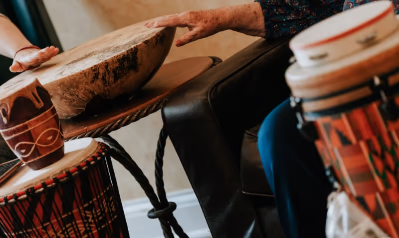 Close-up of two people playing hand drums indoors, showing their hands and parts of the drums with colorful patterns.