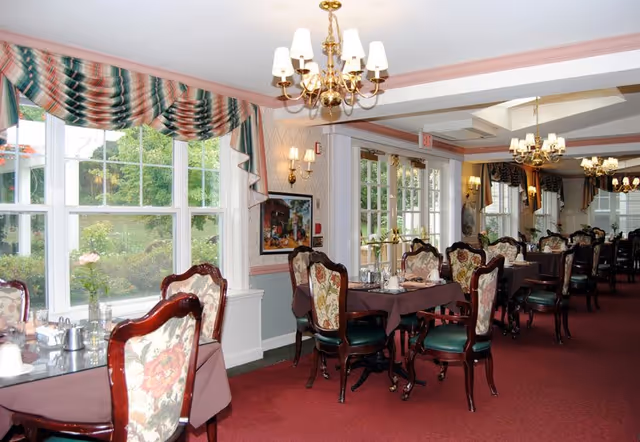 Elegant dining room with multiple set tables and upholstered chairs, chandeliers, and large windows with patterned valances overlooking greenery.