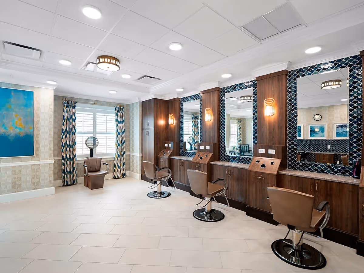 Interior view of a salon area in an assisted living facility featuring three brown salon chairs in front of large mirrors with wooden frames and blue patterned tile accents. The room has light-colored tiled flooring, a window with patterned curtains, a hair dryer chair, and modern ceiling lights.