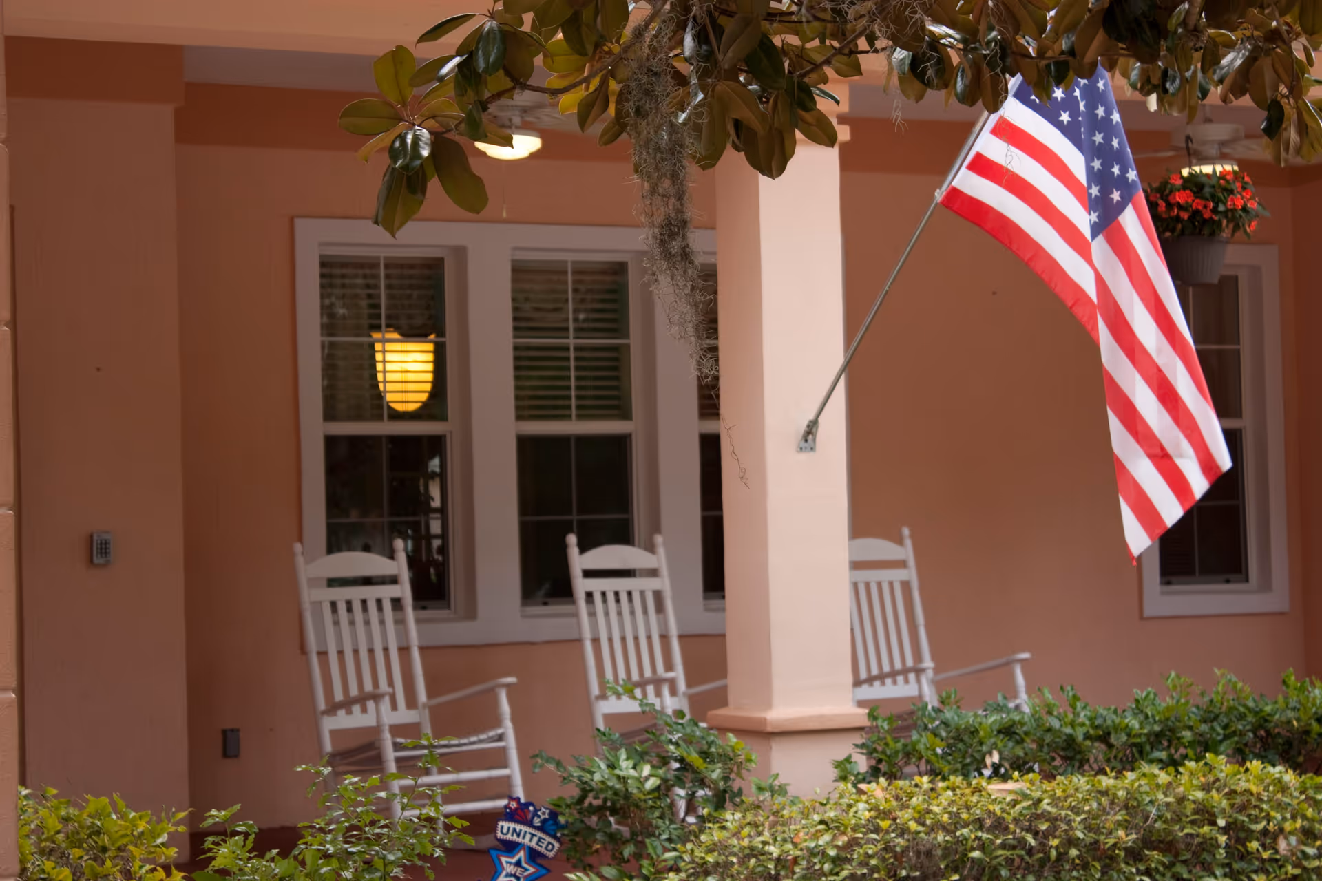 Front porch of a building with peach-colored walls, three white rocking chairs, an American flag mounted on a pillar, green shrubs in the foreground, and a hanging flower pot with red flowers.