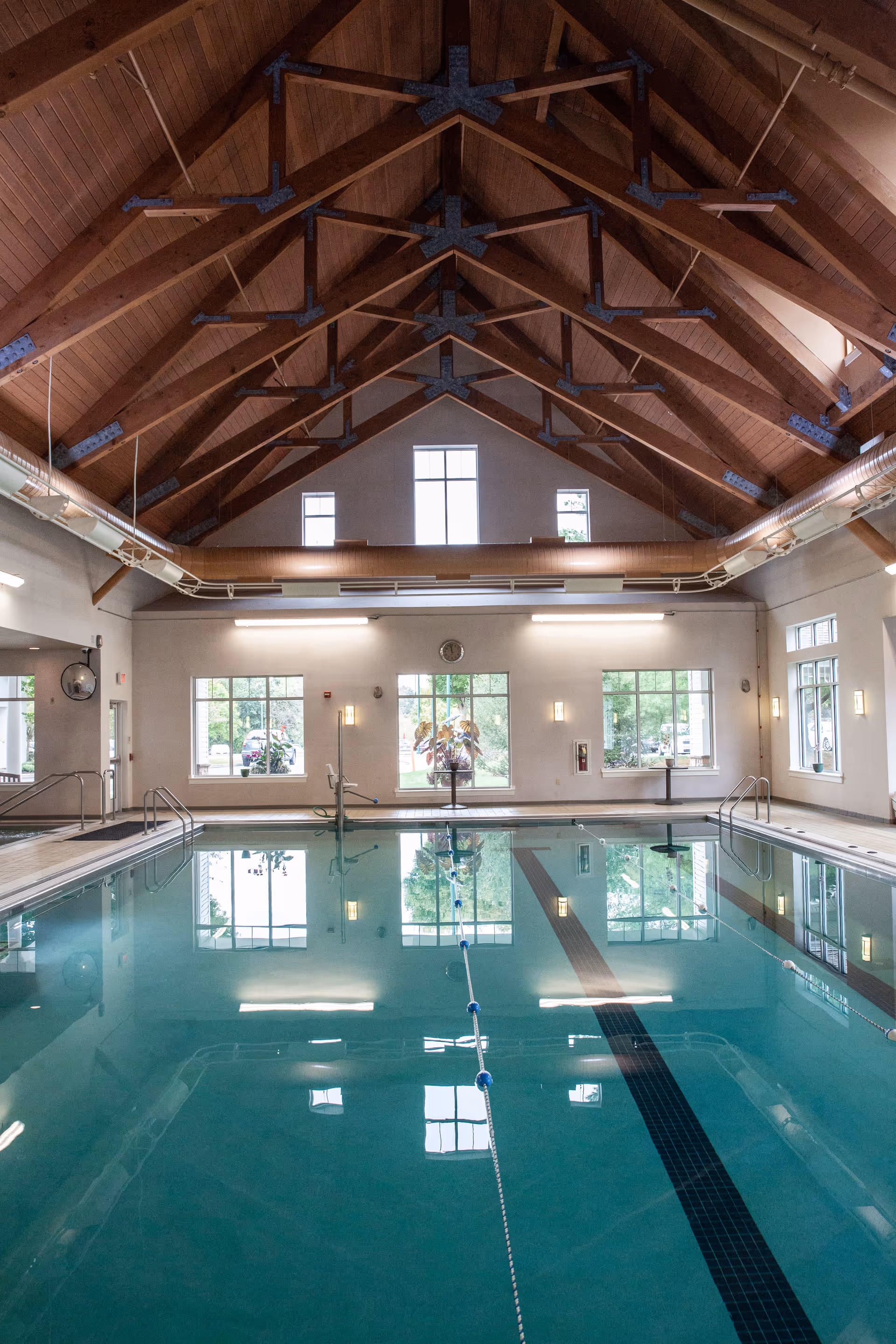 Indoor swimming pool in a high-ceilinged room with wooden trusses and large windows reflected in the water.