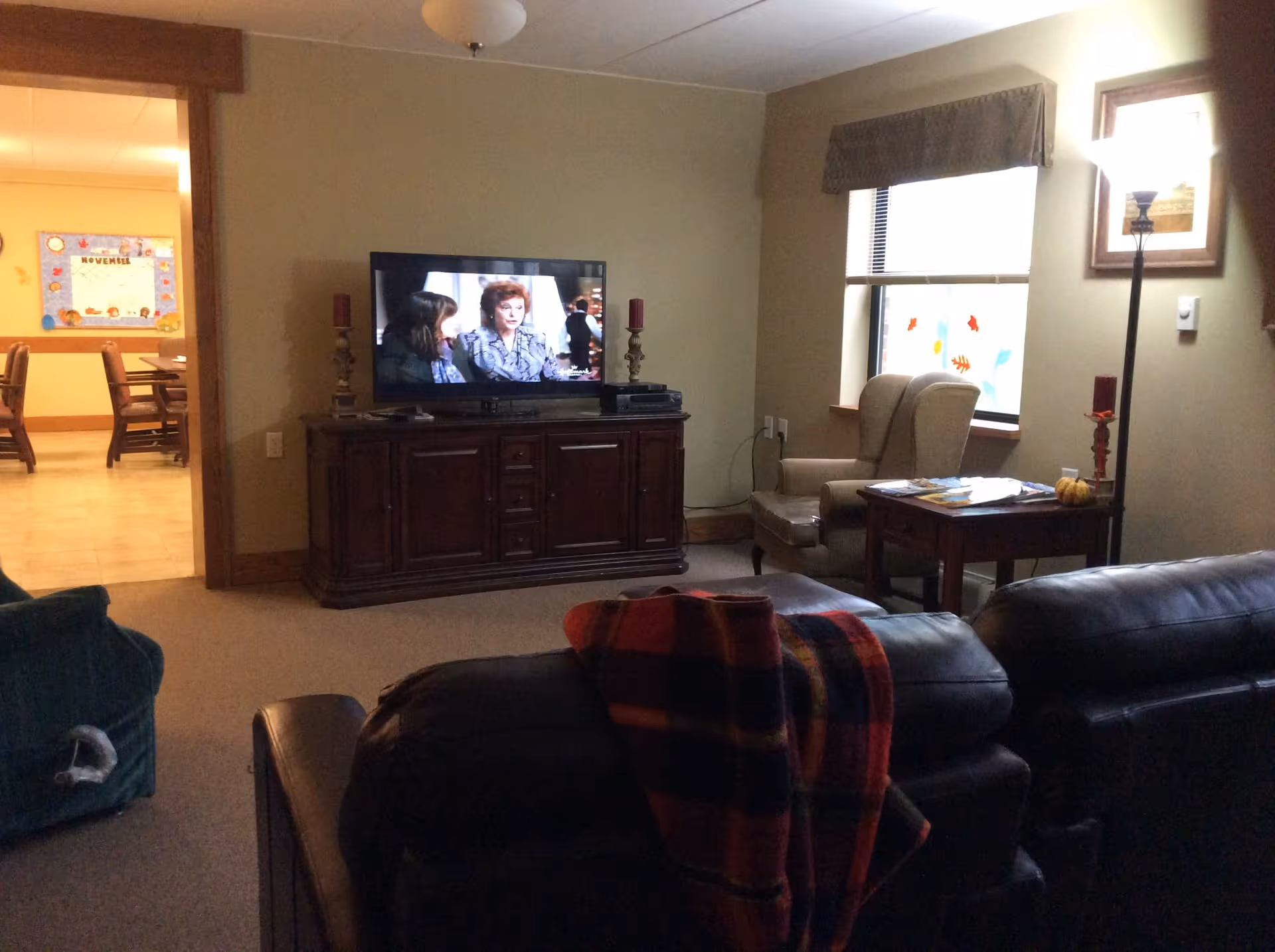 A cozy living room with a dark leather couch in the foreground, a wooden TV stand with a flat-screen television showing a scene with two women, a beige armchair next to a window with a valance, a wooden side table with magazines and a small pumpkin, and a floor lamp. In the background, there is a dining area with wooden chairs and a bulletin board on the wall.