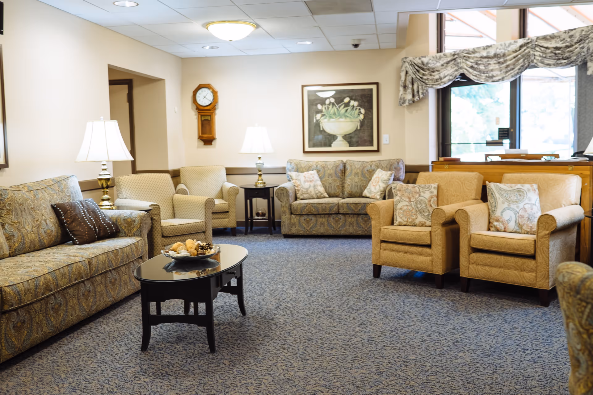 A cozy living room area with patterned sofas and armchairs arranged around a black coffee table. The room has two table lamps, a wall clock, a framed floral painting, and a window with patterned valance curtains letting in natural light.