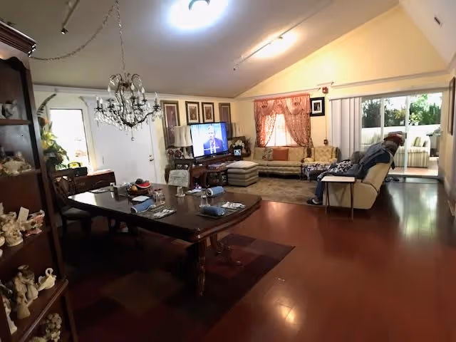 A spacious living room with wooden flooring featuring a large dining table set with plates and napkins on the left side. A chandelier hangs above the dining table. In the background, there is a seating area with a television showing a person speaking, a beige sectional sofa with cushions, and a window with pink curtains. A sliding glass door leads to an outdoor patio area with additional seating.