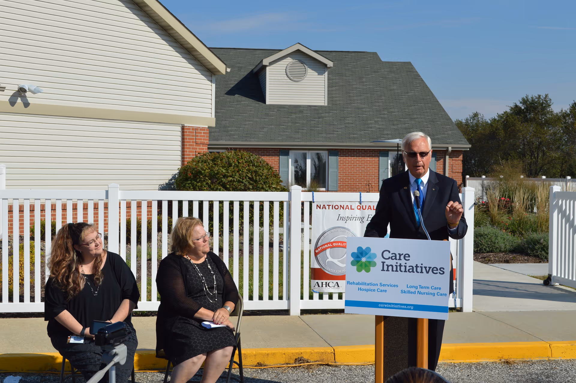 An outdoor event at Atlantic Specialty Care with a man in a suit and sunglasses speaking at a podium labeled Care Initiatives. Two women are seated to his left, listening attentively. Behind them is a white fence and a building with brick and siding exterior under a clear blue sky.