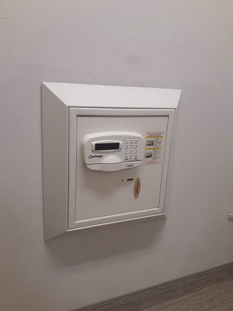 A white wall-mounted safe with a digital keypad and a small display screen, labeled 'Challenger', installed in a light-colored wall above a carpeted floor.
