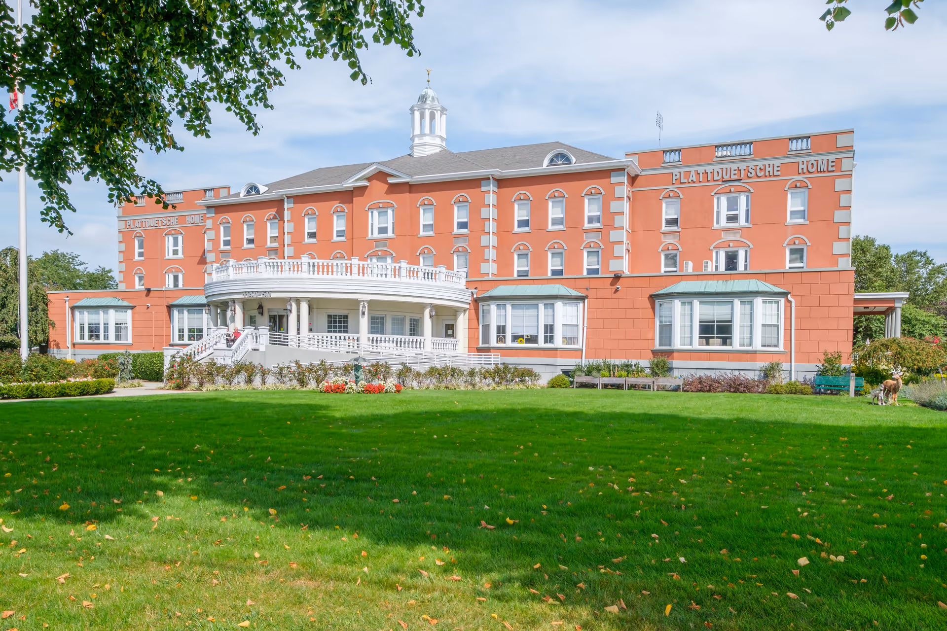 Exterior view of the Plattduetsche Home Society building, a large multi-story brick structure with white trim and a cupola on the roof. The building features a prominent rounded porch with white railings and columns. In front of the building is a well-maintained green lawn with some flower beds and trees framing the scene.