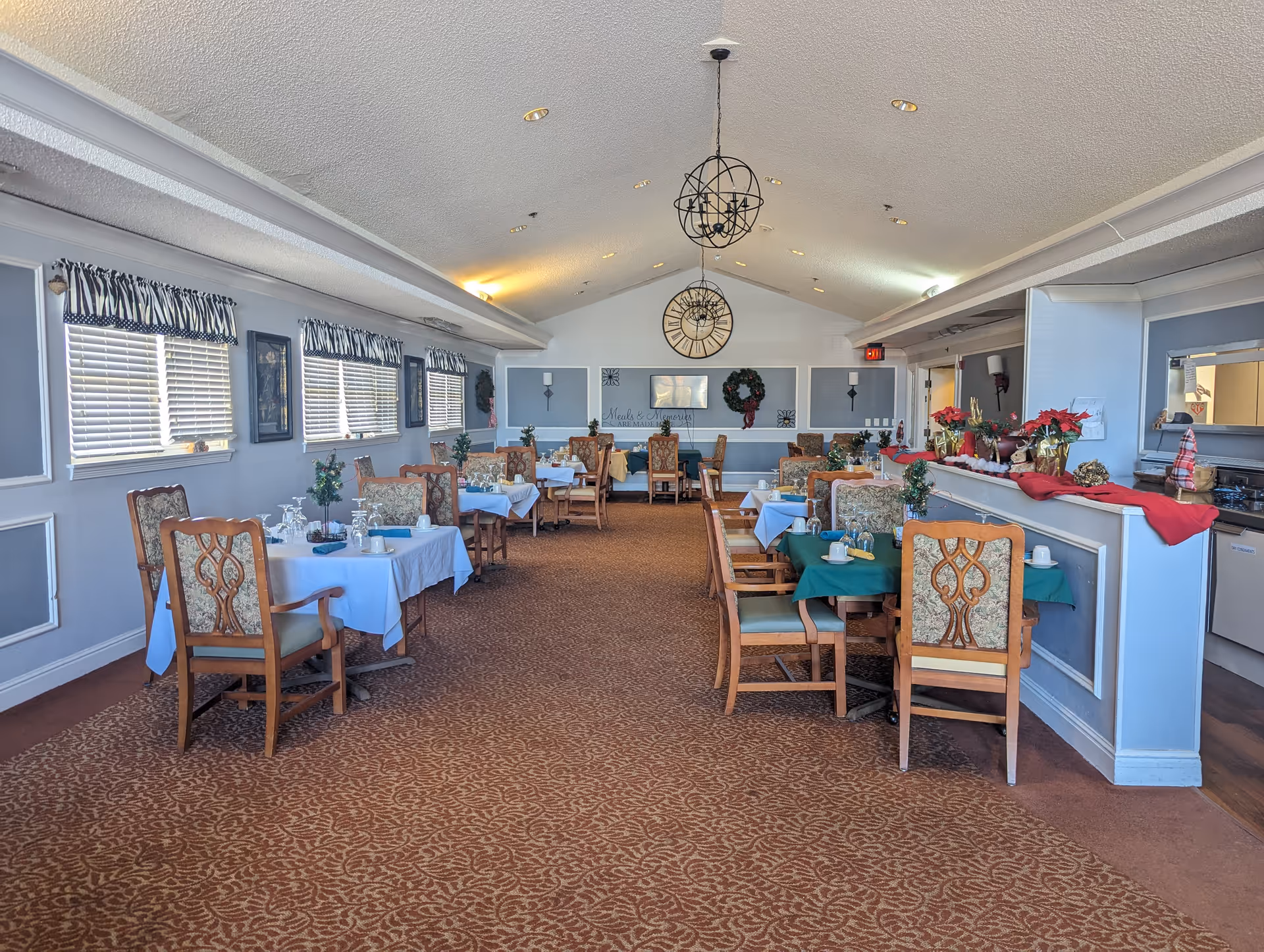 A bright dining room with multiple set tables and wooden chairs, decorative pendant lights, and holiday wreaths and poinsettias.