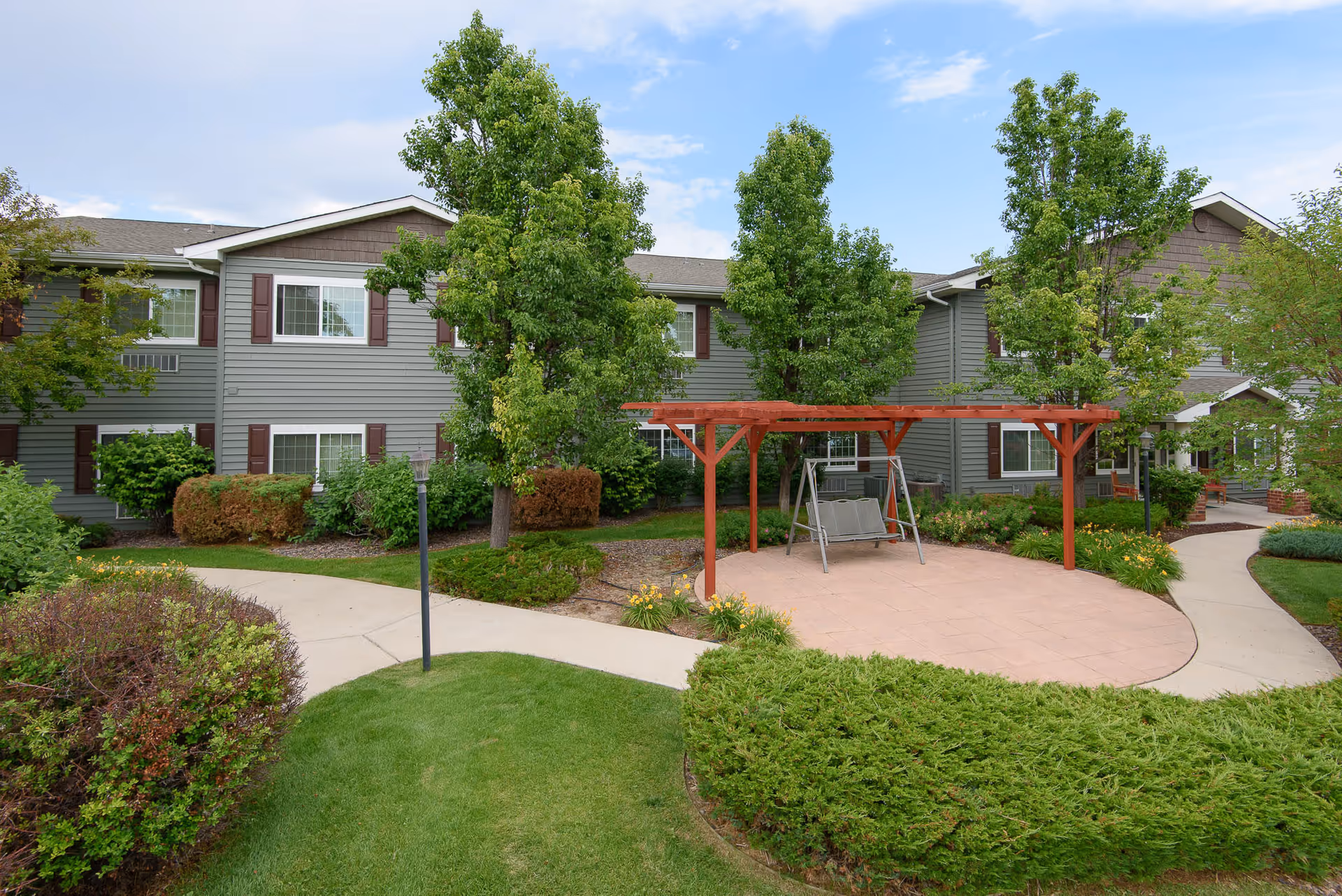 Courtyard with a pergola and bench swing in front of a two-story gray residential building surrounded by trees and landscaped pathways.