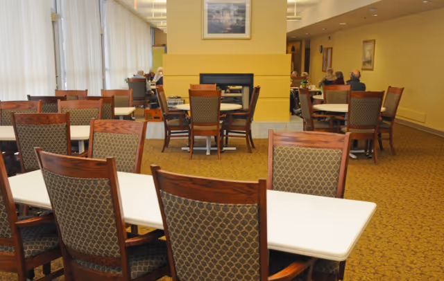 Interior view of a senior living facility dining area with multiple tables and chairs arranged neatly. Several elderly people are seated at tables in the background near a yellow wall with a fireplace and framed artwork above it. The room has carpeted flooring and large windows with sheer curtains on the left side.