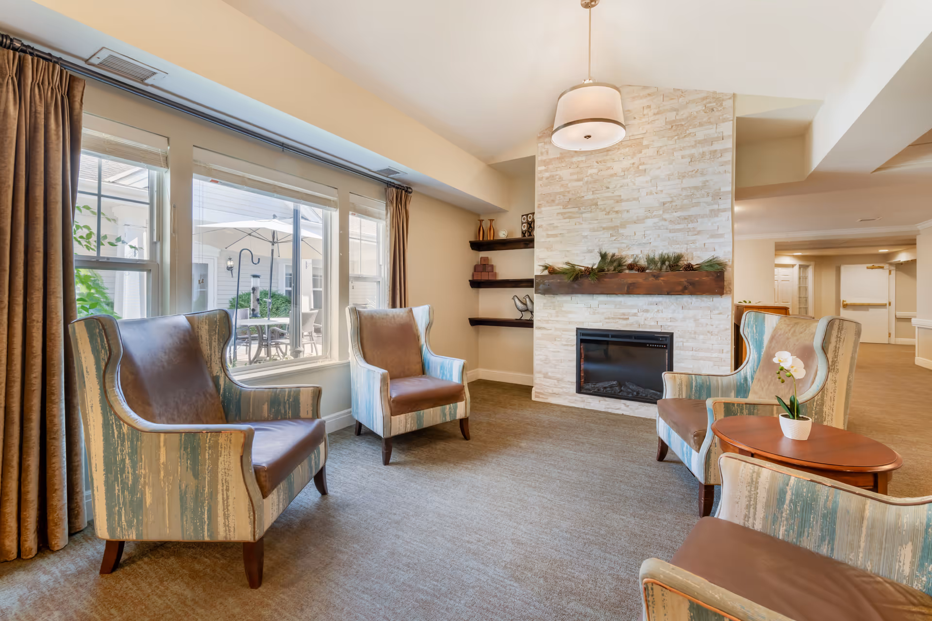 A cozy sitting area in a senior living facility with four armchairs arranged around a small wooden table with a potted orchid. The room features a modern fireplace with a stone surround and a wooden mantel decorated with greenery. Large windows with brown curtains let in natural light and offer a view of an outdoor patio with tables and umbrellas.