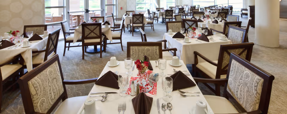 A spacious dining room with multiple tables set for a meal. Each table is covered with a white tablecloth, brown folded napkins, glassware, and silverware. The chairs have patterned upholstery and wooden frames. Large windows in the background allow natural light to fill the room.