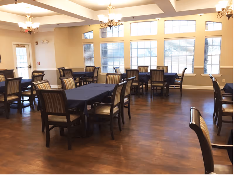 A spacious dining room with multiple tables covered in dark tablecloths and surrounded by wooden chairs with striped cushions. Large windows allow natural light to fill the room, and elegant chandeliers hang from the ceiling.