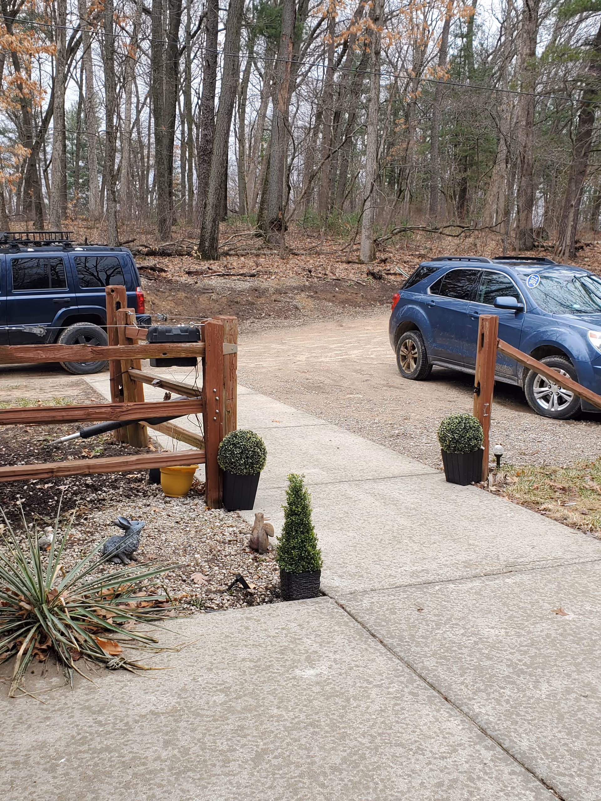 Outdoor view of a parking area with two parked cars near a wooden fence and a concrete walkway. There are small potted plants and garden decorations along the edge of the walkway, with leafless trees in the background.