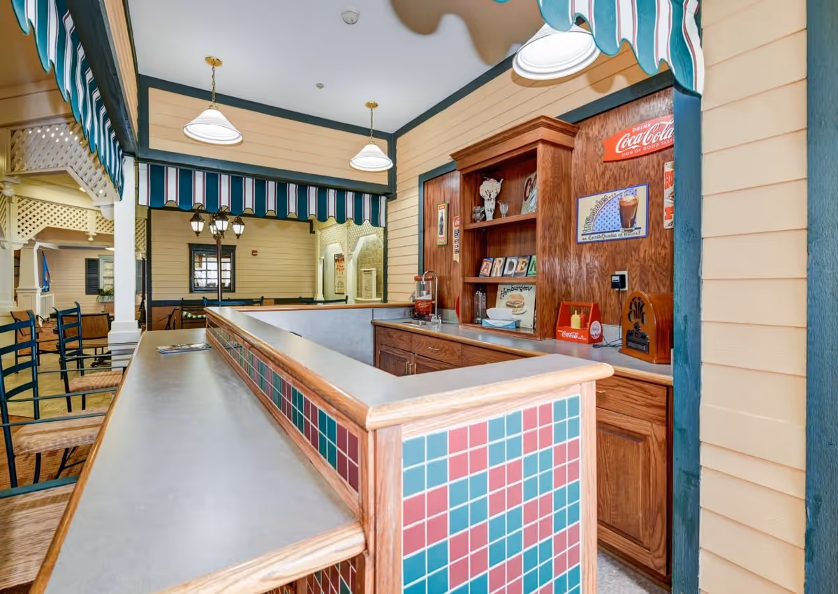 Interior view of a retro-style snack bar or soda fountain area with a counter featuring red and green tiles, wooden cabinetry, and vintage Coca-Cola signs on the wall. The space has striped awnings and hanging pendant lights, with seating visible in the background.