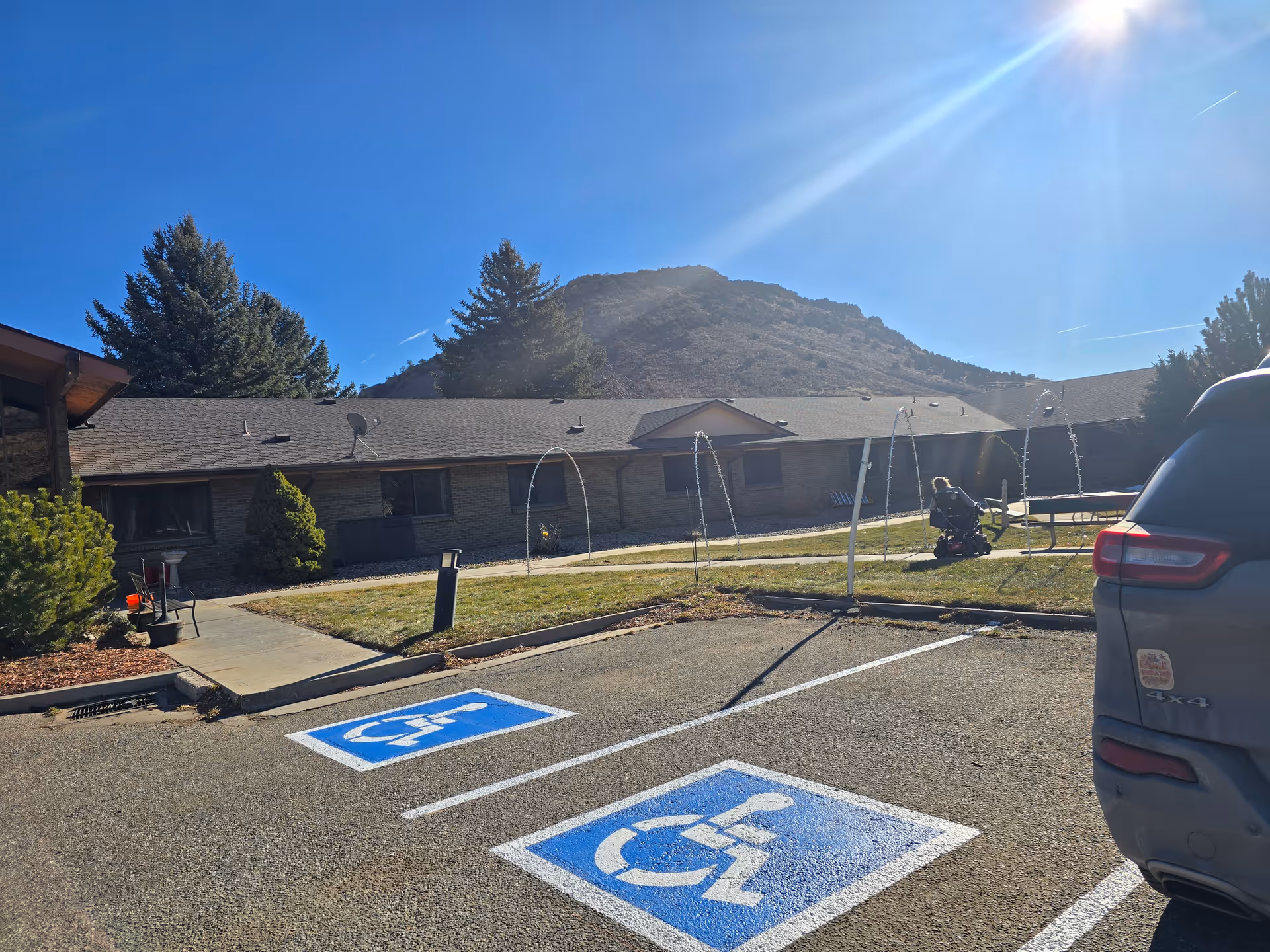Parking lot with two handicap spaces in front of a one-story brick care facility, a person on a mobility scooter on the lawn, and a mountain in the background.