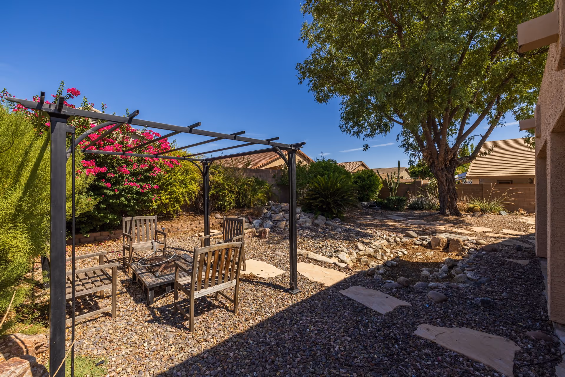 Outdoor seating area with four wooden chairs arranged around a square fire pit under a black metal pergola, surrounded by gravel and landscaping including bushes, a large tree, and flowering plants under a clear blue sky.
