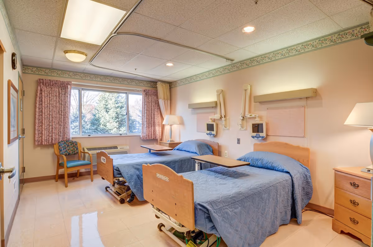 A bright shared patient bedroom with two hospital-style beds with blue bedspreads, bedside tables and wall-mounted medical equipment, a window with curtains and a chair.